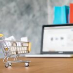 Wózek zakupowy z paczkami ustawiony przed laptopem jako metafora sprzedaży na platformach typu marketplace