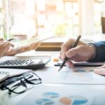 Zbliżenie na biurko księgowe, ręka księgowego zaznaczająca dane na dokumentach, komputer i akcesoria używane przy rozliczeniach estońskiego CIT